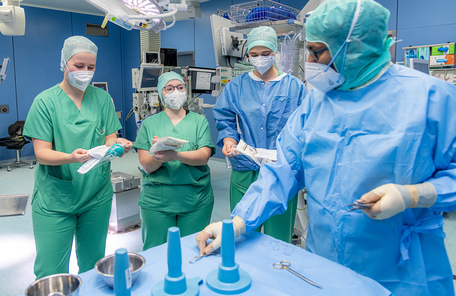 Operationsteam in einem OP-Saal bei der Vorbereitung chirurgischer Instrumente, drei Personen in gr&uuml;ner und eine Person in blauer OP-Kleidung und Maske.